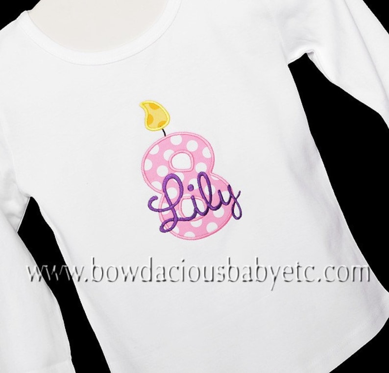 May include: White long-sleeved shirt with a pink number eight with white polka dots and a yellow flame on top. The number eight has the name "Lily" written in purple cursive.