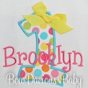 May include: A white onesie with a blue outlined number one with colorful polka dots. A yellow bow is on top of the number. The name "Brooklyn" is embroidered in pink below the number. The text "Bow Dacious Baby" is embroidered in white below the name.