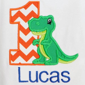 Könnte beinhalten: Ein weißer Strampler mit einem grünen Dinosaurier und einer großen orange-weißen Chevron-Zahl eins. Der Strampler ist mit dem Namen "Lucas" in Blau personalisiert.