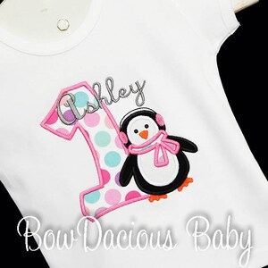 May include: White short-sleeved shirt with a colorful embroidered design. Features a large number 1 with a polka dot pattern and the name "Ashley" above a penguin wearing earmuffs and a scarf. The text "Bow Dacious Baby" is at the bottom.