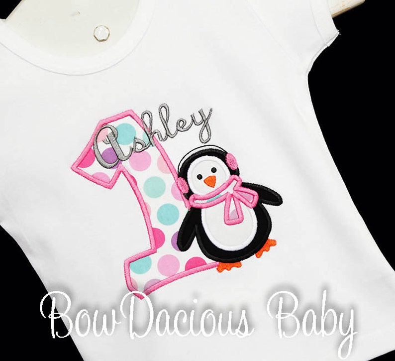 May include: White short-sleeved shirt with a colorful embroidered design. Features a large number 1 with a polka dot pattern and the name "Ashley" above a penguin wearing earmuffs and a scarf. The text "Bow Dacious Baby" is at the bottom.