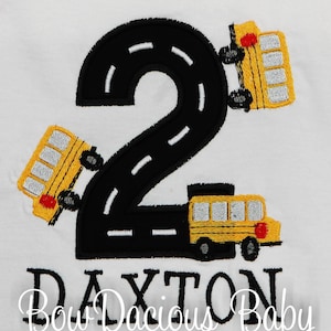 May include: A white onesie with a black number 2 shaped like a road with three yellow school buses driving on it. The text "Bow Dacious Baby" and the name "DAYTON" are embroidered on the onesie.