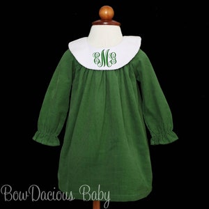 May include: A green corduroy dress with long sleeves and a white Peter Pan collar. The collar has a green embroidered monogram. The dress features ruffled cuffs and is displayed on a mannequin. The text "Bow Dacious Baby" is at the bottom.