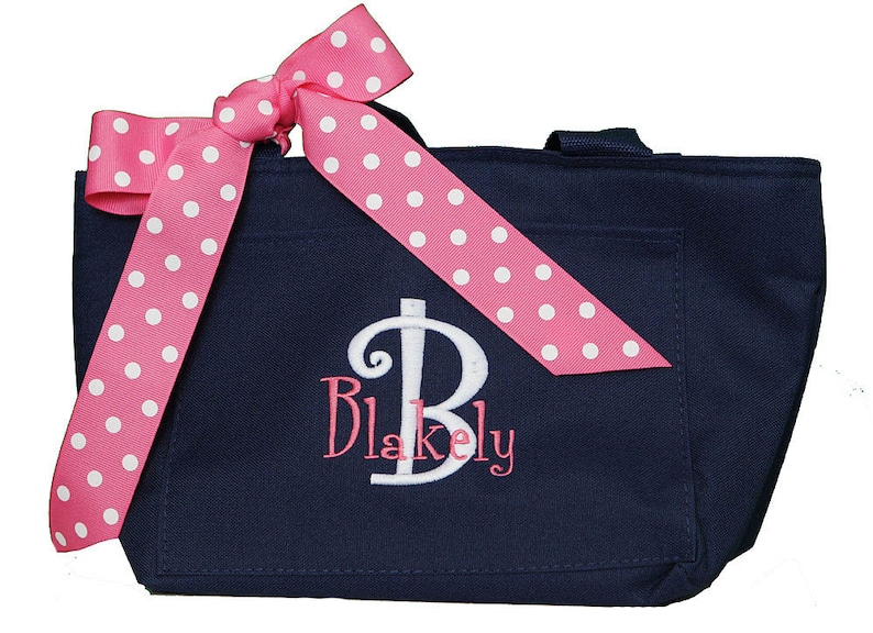 K&ouml;nnte beinhalten: Eine marineblaue Tragetasche mit einer Schleife aus rosa Band mit wei&szlig;en Punkten. Die Tasche hat ein wei&szlig; gesticktes Monogramm mit der Aufschrift "Blakely".