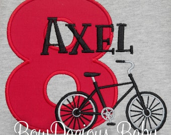May include: A gray t-shirt with a red number 8 and the name "AXEL" embroidered in black. A black bicycle with white accents is positioned next to the number. The text "Bow Dacious Baby" is at the bottom.