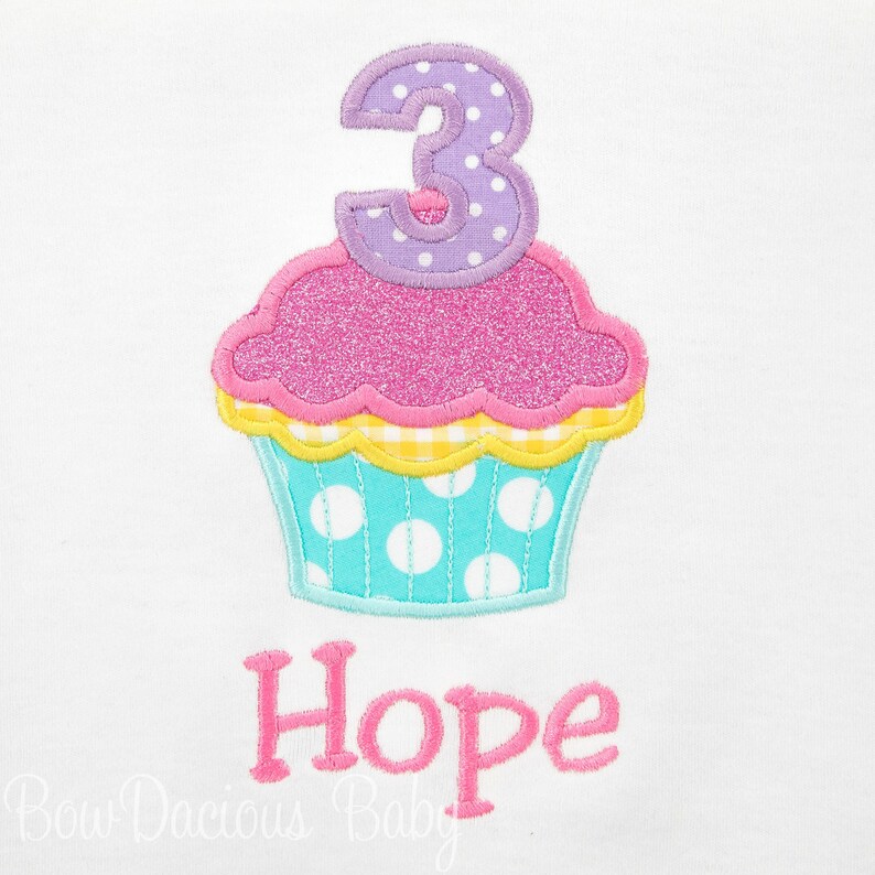 May include: A white onesie with a pink and blue cupcake design and the number 3 on top. The cupcake has yellow and white checked frosting and white polka dots on the bottom. The name "Hope" is embroidered below the cupcake.