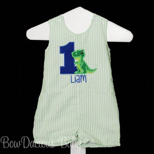 May include: Green and blue striped baby onesie with a blue number one and a green dinosaur. The onesie is personalized with the name "Liam".