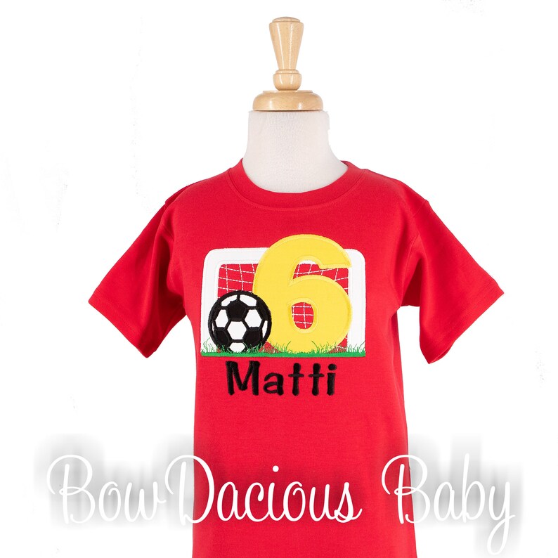 May include: Red short-sleeved t-shirt with a soccer design. The design features a soccer ball, a yellow number 6, and the name "Matti" in black. The shirt is displayed on a mannequin.