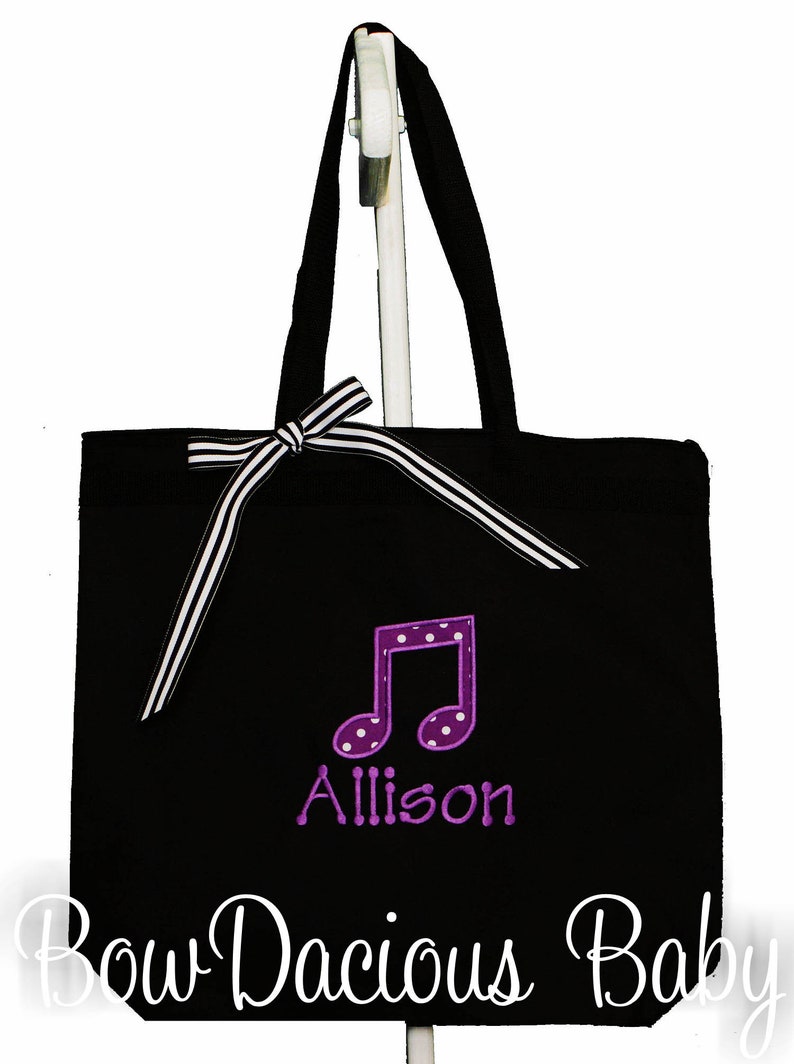May include: Black canvas tote bag with a white and black striped ribbon bow. The bag has a purple musical note with white polka dots and the name "Allison" embroidered on it. The text "Bow Dacious Baby" is printed on the bottom of the bag.