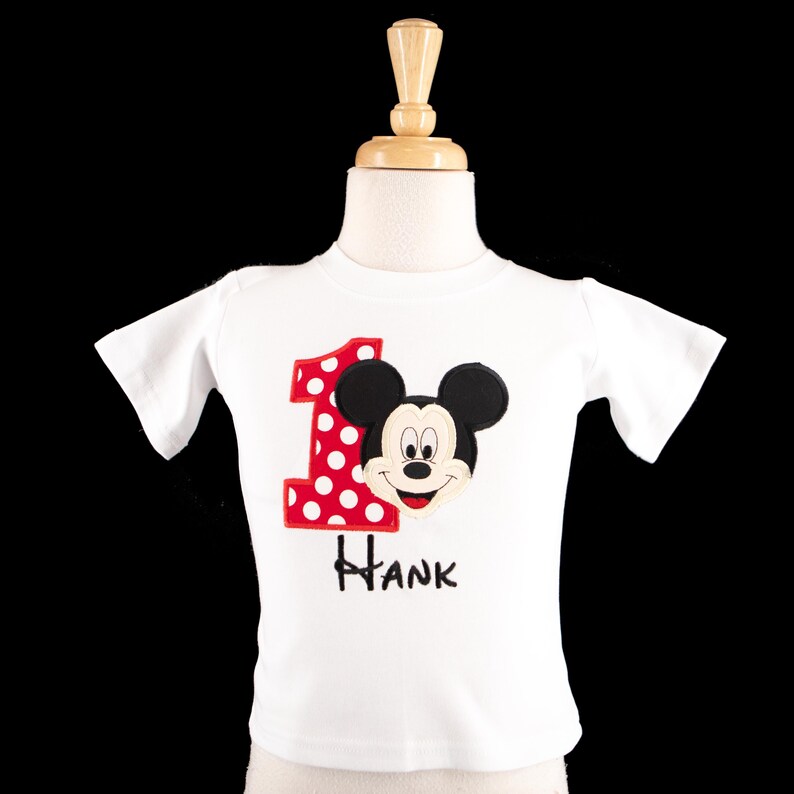May include: White short-sleeved t-shirt featuring a Mickey Mouse design. The number "1" in red with white polka dots is on the left, and the name "Hank" is printed below the character's face. The shirt is displayed on a mannequin.