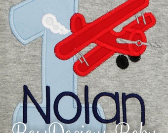 May include: A blue number one with a red airplane flying over it. The name Nolan is written in navy blue below the number. The words "Bow Dacious Baby" are written in white below the name.