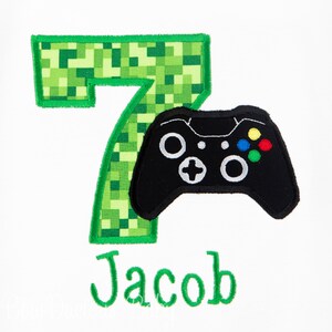 May include: Green pixelated number seven with a black video game controller on top. The name Jacob is written below in green.