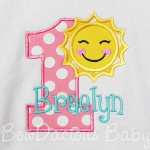 K&ouml;nnte beinhalten: Eine rosa Eins mit wei&szlig;en Punkten, eine l&auml;chelnde gelbe Sonne und der Name "Braelyn" in t&uuml;rkisfarbener Stickerei. Das Design befindet sich auf wei&szlig;em Hintergrund, ideal f&uuml;r den ersten Geburtstag.