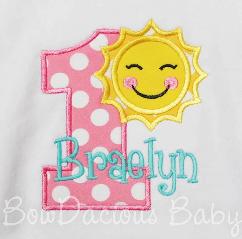 K&ouml;nnte beinhalten: Eine rosa Eins mit wei&szlig;en Punkten, eine l&auml;chelnde gelbe Sonne und der Name "Braelyn" in t&uuml;rkisfarbener Stickerei. Das Design befindet sich auf wei&szlig;em Hintergrund, ideal f&uuml;r den ersten Geburtstag.
