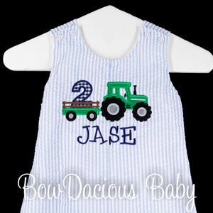 May include: A blue and white striped seersucker romper with a green tractor and wagon applique. The number "2" is featured, along with the name "JASE" in navy blue. The romper is displayed on a white hanger.