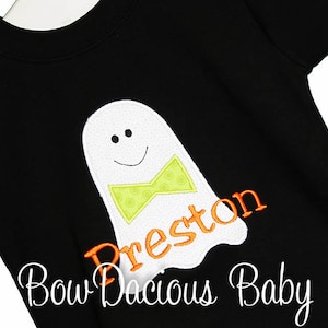 May include: Black short-sleeved shirt featuring a white ghost applique with a green bow tie and the name "Preston" in orange. The ghost has a smiling face. The text "Bow Dacious Baby" is written in cursive at the bottom.