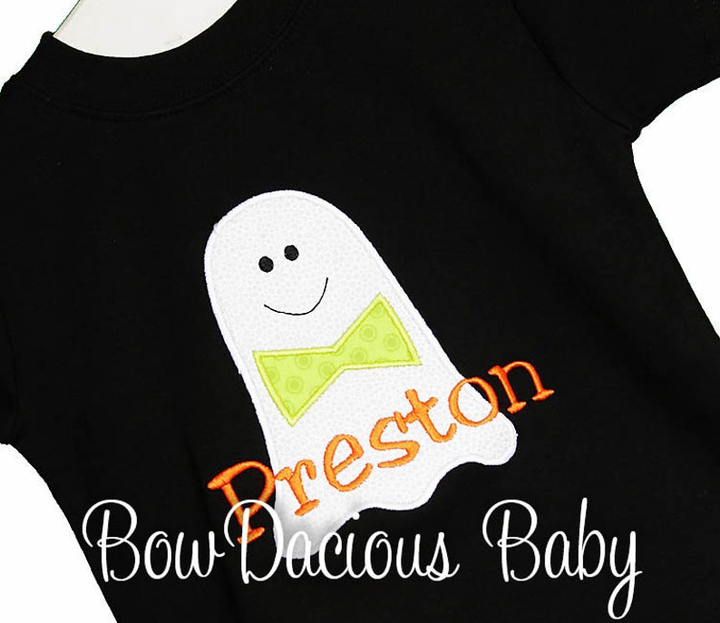 May include: Black short-sleeved shirt featuring a white ghost applique with a green bow tie and the name "Preston" in orange. The ghost has a smiling face. The text "Bow Dacious Baby" is written in cursive at the bottom.