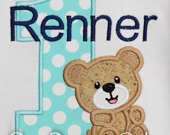 May include: A turquoise blue and white polka dot number one with a brown teddy bear to the right. The name "Renner" is embroidered in navy blue above the number. The text "Bow Dacious Baby" is embroidered in white below the number.