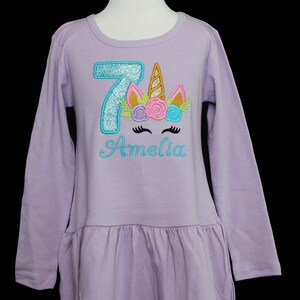 Puede incluir: Una camisa de manga larga lavanda con un n&uacute;mero 7 azul brillante y una cara de unicornio con un cuerno dorado y flores rosas y azules. La camisa dice "Amelia" en letras azules.