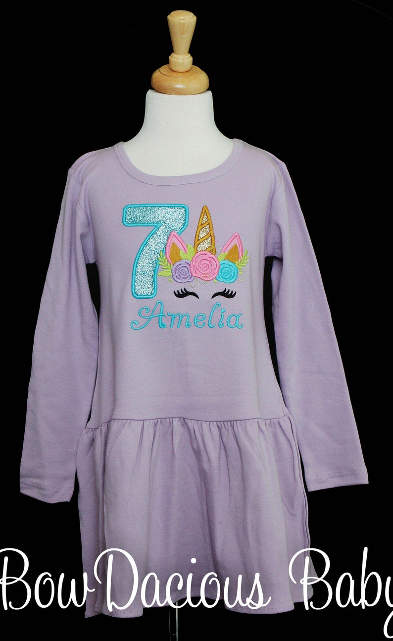 Puede incluir: Una camisa de manga larga lavanda con un n&uacute;mero 7 azul brillante y una cara de unicornio con un cuerno dorado y flores rosas y azules. La camisa dice "Amelia" en letras azules.