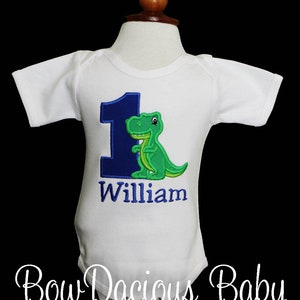 May include: White baby bodysuit with a blue number 1 and a green dinosaur applique. The name "William" is embroidered below the number. The bodysuit is displayed on a mannequin.