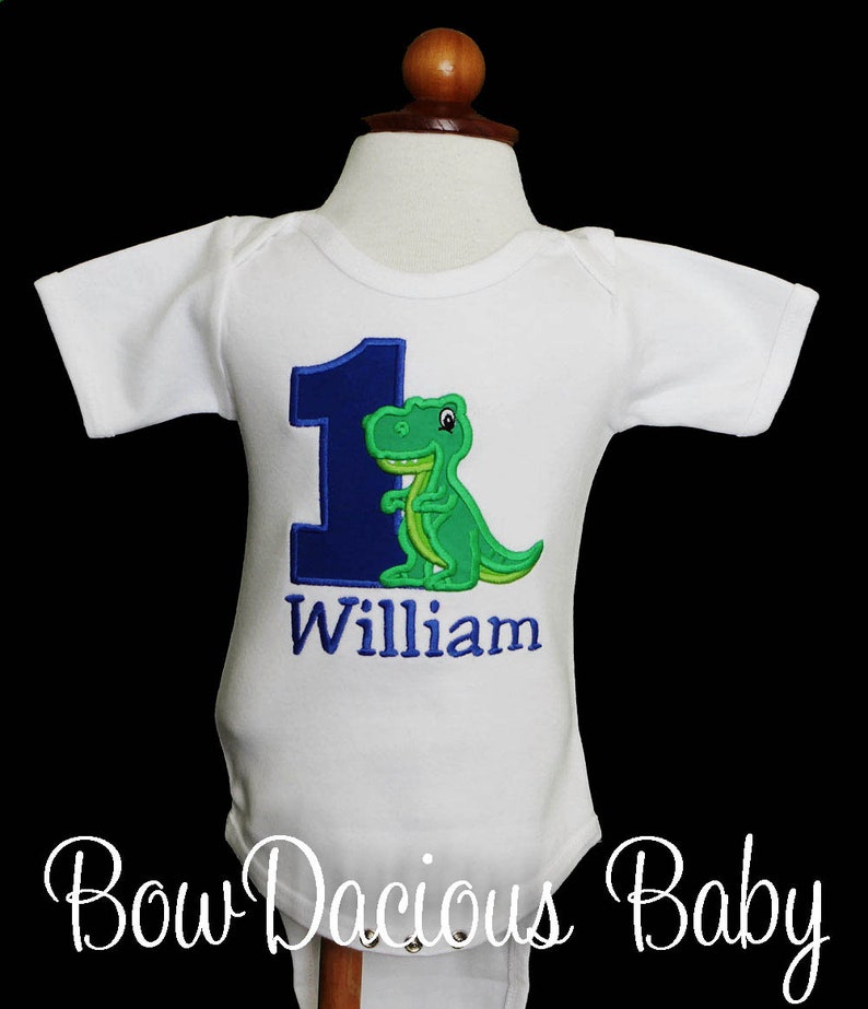 May include: White baby bodysuit with a blue number 1 and a green dinosaur applique. The name "William" is embroidered below the number. The bodysuit is displayed on a mannequin.