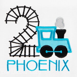 Könnte beinhalten: Ein blauer und weißer Zug mit einer schwarzen Schiene in Form der Zahl zwei. Der Zug hat einen schwarzen Schornstein und eine graue Rauchwolke. Das Wort "PHOENIX" ist in Blau unter dem Zug gestickt.