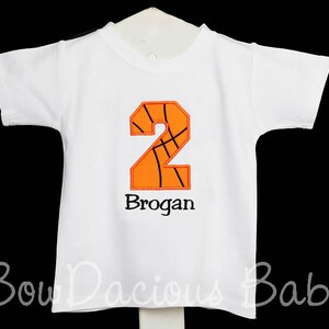 May include: White short-sleeved t-shirt with an orange and black basketball-themed number 2 applique. The name "Brogan" is embroidered below the number. The shirt is designed for a child.