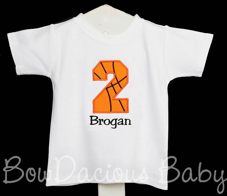 May include: White short-sleeved t-shirt with an orange and black basketball-themed number 2 applique. The name "Brogan" is embroidered below the number. The shirt is designed for a child.