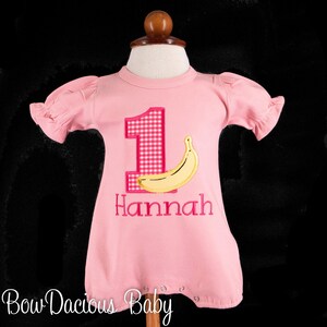 Könnte beinhalten: Rosa Baby-Strampler mit kurzen Rüschenärmeln. Die Zahl "1" ist mit einem rosa Vichy-Muster appliziert, daneben eine gelbe Banane. Der Name "Hannah" ist darunter gestickt. Die Worte "Bow Dacious Baby" stehen unten.
