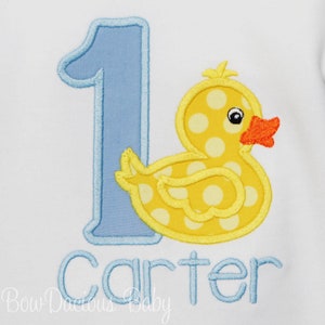 May include: A blue number one with a yellow rubber duck with white polka dots and an orange beak. The name Carter is written in blue below the number and duck.