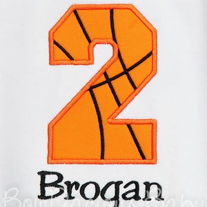 May include: An orange number 2, designed to resemble a basketball, with black lines and a black outline. Below the number is the name "Brogan" in black lettering, embroidered on a white background.