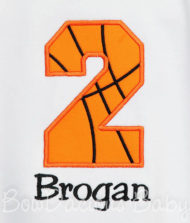 May include: An orange number 2, designed to resemble a basketball, with black lines and a black outline. Below the number is the name "Brogan" in black lettering, embroidered on a white background.