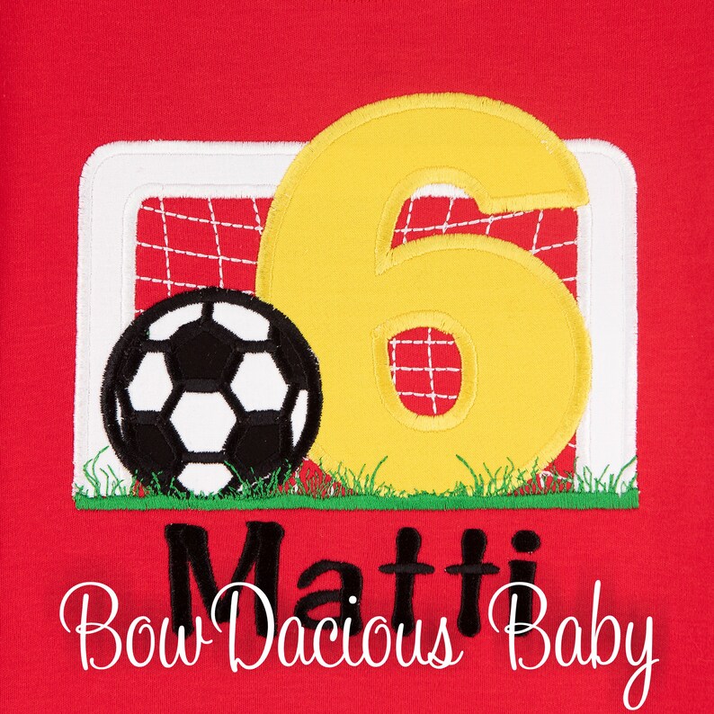 May include: Red shirt with a football theme. Features a black and white football, a yellow number 6, and a goal. The name "Matti" and the text "Bow Dacious Baby" are also displayed.