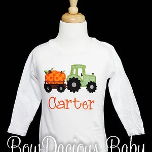 May include: White long-sleeved shirt featuring an embroidered green tractor pulling a wagon with two orange pumpkins. The name "Carter" is embroidered in orange. The shirt is displayed on a mannequin.