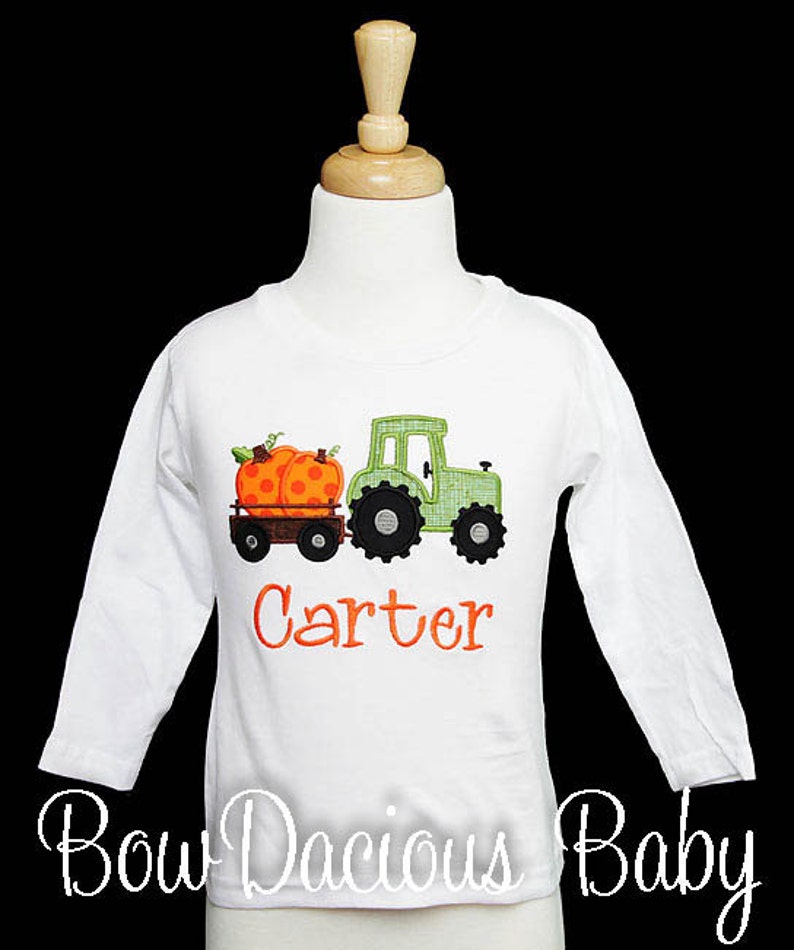 May include: White long-sleeved shirt featuring an embroidered green tractor pulling a wagon with two orange pumpkins. The name "Carter" is embroidered in orange. The shirt is displayed on a mannequin.