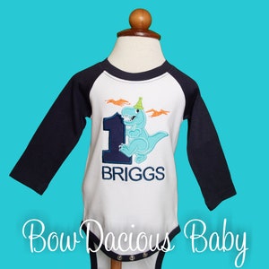 May include: A white and navy blue baby bodysuit with long sleeves, featuring a light blue dinosaur wearing a party hat and the number 1. The name "BRIGGS" is embroidered below. The text "Bow Dacious Baby" is at the bottom of the bodysuit.