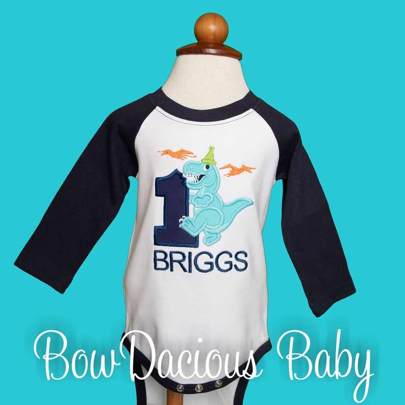 May include: A white and navy blue baby bodysuit with long sleeves, featuring a light blue dinosaur wearing a party hat and the number 1. The name "BRIGGS" is embroidered below. The text "Bow Dacious Baby" is at the bottom of the bodysuit.