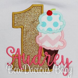 Könnte beinhalten: Goldene Glitzerzahl eins mit einem rosa, weißen und blauen Eiswaffel mit einer Kirsche oben drauf. Der Text "Audrey Bow Dacious Baby" befindet sich unter der Zahl.