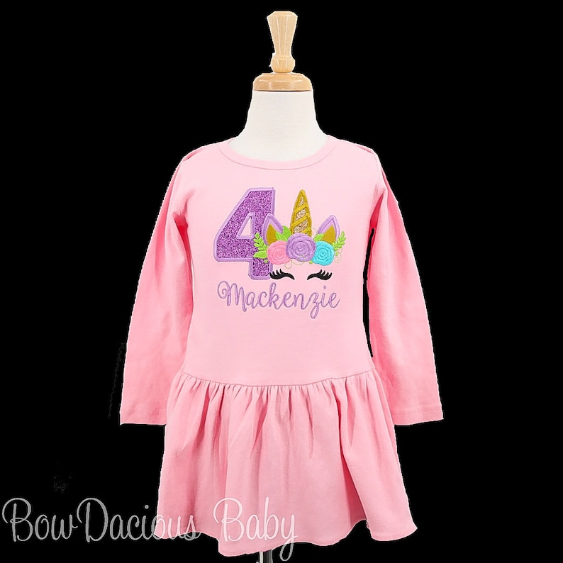 Puede incluir: Vestido rosa de manga larga con un n&uacute;mero 4 de purpurina morada y una cara de unicornio con flores y un cuerno dorado. El vestido dice "Mackenzie" debajo del unicornio.