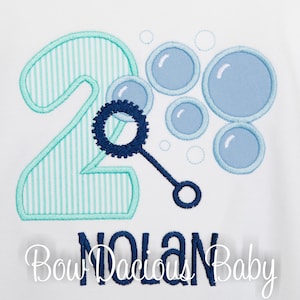 May include: A light blue and green number two with white stripes, a blue bubble wand, and blue bubbles. The text "Nolan" is written below the number. The text "Bow Dacious Baby" is written below the name.