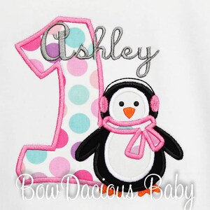 May include: White t-shirt with an embroidered design featuring a large number one with pink polka dots and a pink outline. A penguin wearing earmuffs and a scarf is next to the number. The name "Ashley" is above the penguin.
