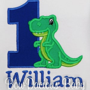 May include: White onesie with a large blue number 1 and a green dinosaur applique. The dinosaur has light green accents. The name "William" is embroidered in blue script below the number and dinosaur. The text "Bow Dacious Baby" is below the name.