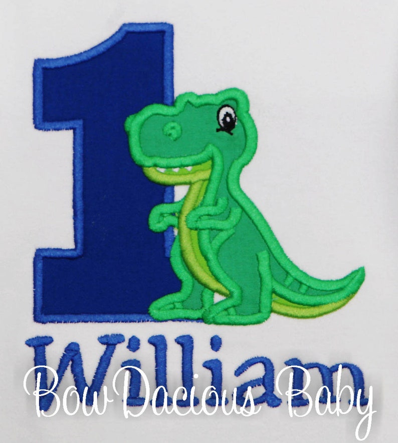 May include: White onesie with a large blue number 1 and a green dinosaur applique. The dinosaur has light green accents. The name "William" is embroidered in blue script below the number and dinosaur. The text "Bow Dacious Baby" is below the name.