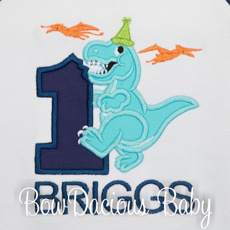 May include: A white shirt with an embroidered design featuring a blue dinosaur wearing a green party hat next to a navy blue number 1. Orange pterodactyls fly in the background. The name "BRIGGS" is embroidered below the number.
