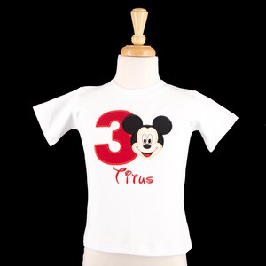 May include: White t-shirt featuring a red number 3 and a Mickey Mouse applique. The shirt also has the name "Cirus" embroidered in red script. The shirt is displayed on a mannequin.