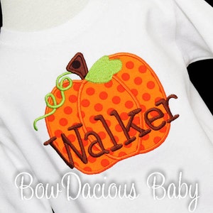 May include: White long-sleeved shirt featuring an orange polka-dotted pumpkin applique with the name "Walker" in brown letters. The pumpkin has a green stem and vine. The text "Bow Dacious Baby" is written in cursive at the bottom.