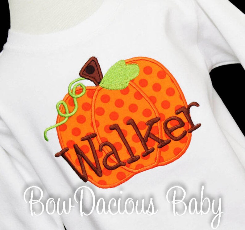 May include: White long-sleeved shirt featuring an orange polka-dotted pumpkin applique with the name "Walker" in brown letters. The pumpkin has a green stem and vine. The text "Bow Dacious Baby" is written in cursive at the bottom.