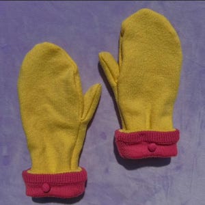 Puede incluir: Un par de mitones amarillos con puños de canalé rosas y un pequeño detalle de botón. Los mitones están hechos de un material suave y están diseñados para mantener las manos calientes en climas fríos.
