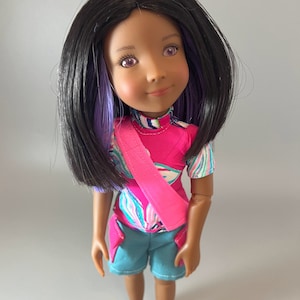 Op de afbeelding: Een pop met bruine huid, zwart haar met paarse strepen en paarse ogen. Ze draagt een roze shirt met een blauw-wit patroon, een blauwe broek en een roze sjerp.